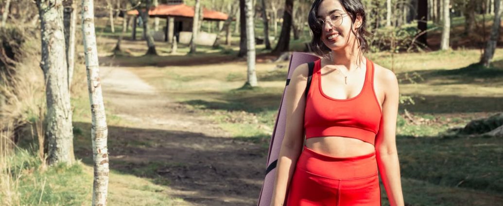 Smiling woman in red sport clothing holding a yoga mat outdoors.