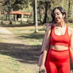 Smiling woman in red sport clothing holding a yoga mat outdoors.